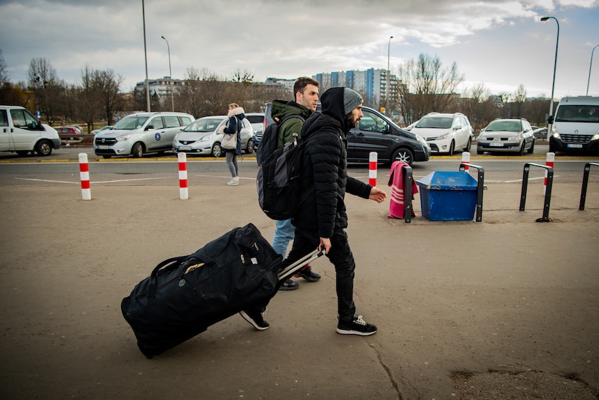 Two men walk with their suitcases across a footpath.