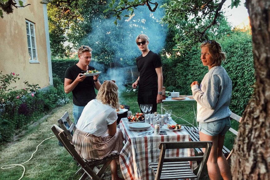 Four young adults, two men and two women, setting up a table with food in a backyard for a party