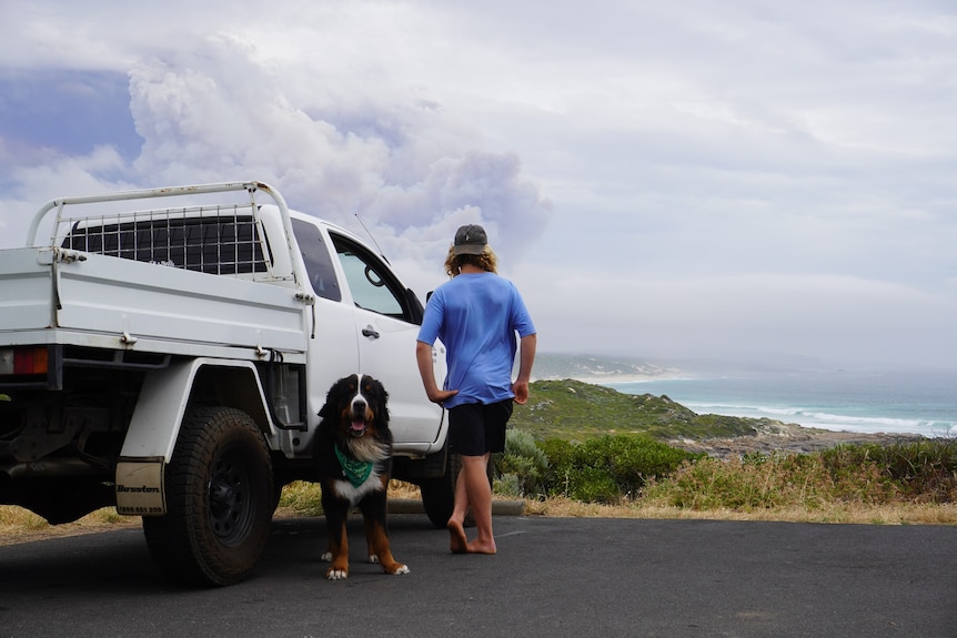 A man with his dog and ute watches smoke billow in the distance