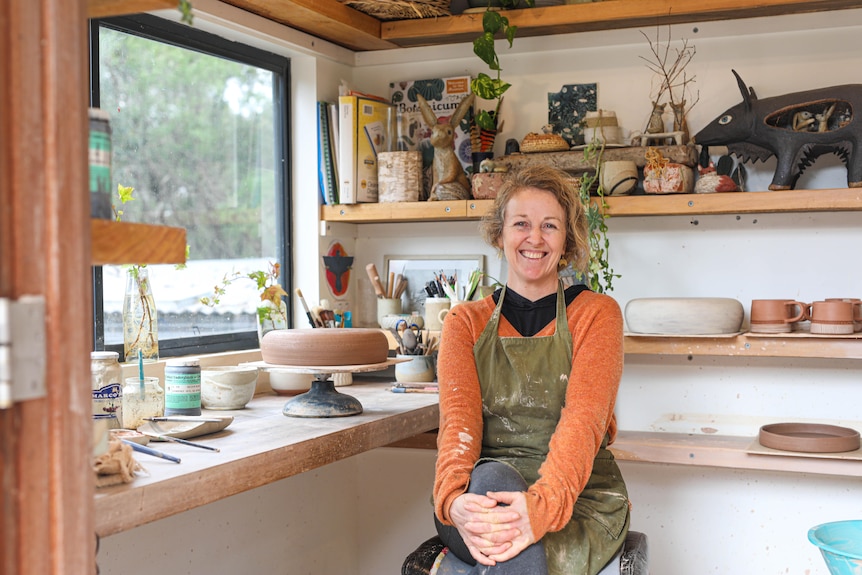 A woman in an orange jumper and green apron has a big smile as she sits in her pottery studio.