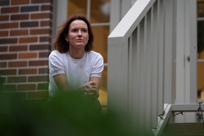 A woman sits on the steps of a balcony of a house, looking ahead with a neutral expression. 