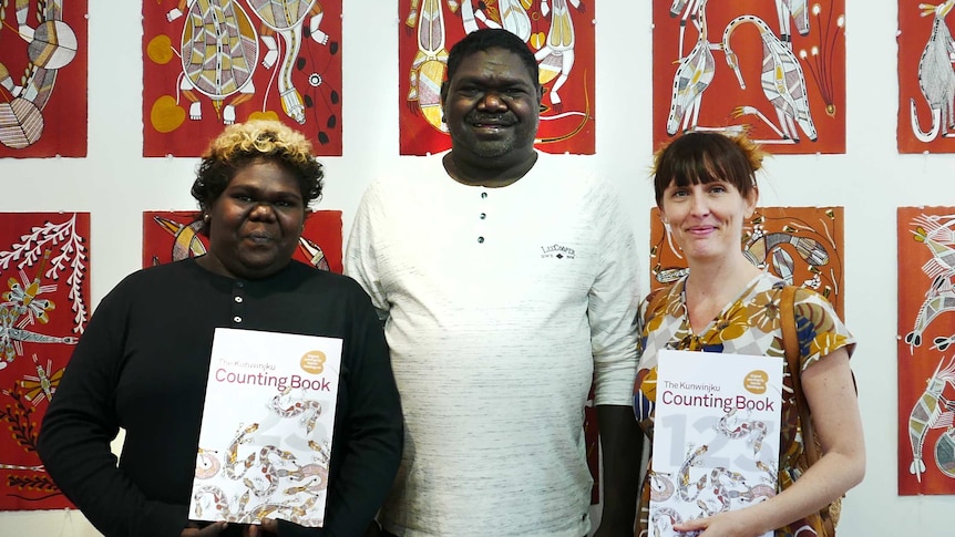 three people holding children's books stand in front of prints of indigenous art