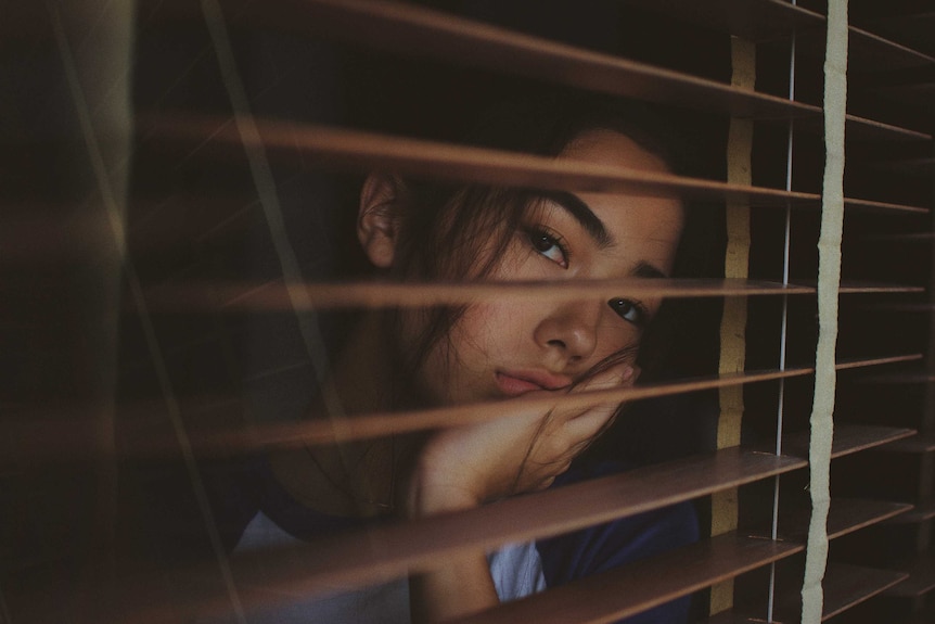 A woman looks despondently through venetian blinds.