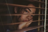 A woman looks despondently through venetian blinds.