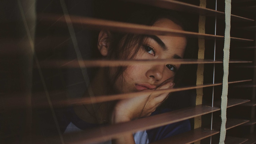 A woman looks despondently through venetian blinds.