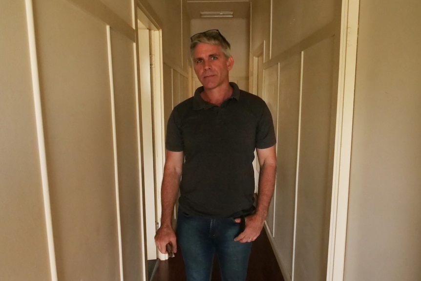 A serious middle-aged man with greying hair, black t-shirt, blue jeans, standing in hallway.