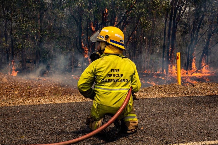 A firefighter kneels on the ground with a hose in front of a bushfire.