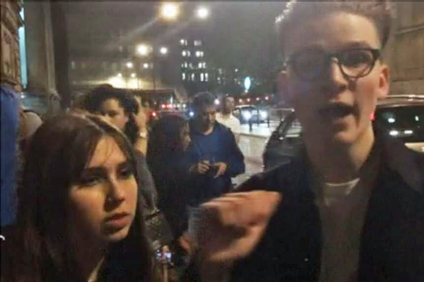 A young man and woman stand among crowd on London street at night.