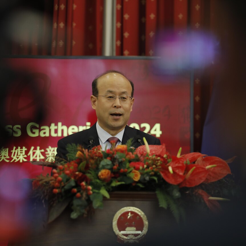 A bald man with glasses speaks at a lectern covered in flowers.