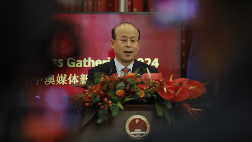 A bald man with glasses speaks at a lectern covered in flowers.