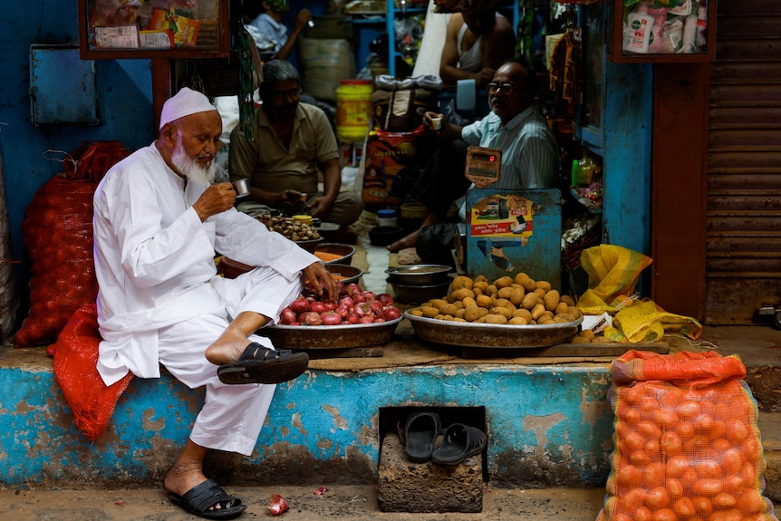 Three shopkeepers take a tea break, sitting near sacks of onions and potatoes in the shade of their shop.