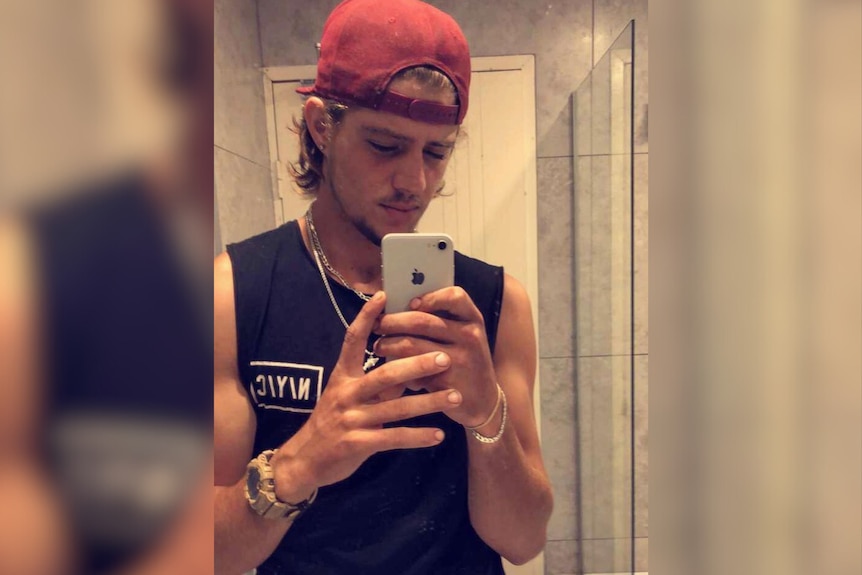 A man in a red cap and a black singlet takes a mirror selfie, he is wearing a chain.