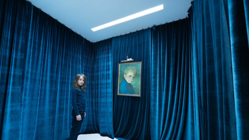 A young boy sits in a blue curtained room with a portrait of himself as a younger boy.