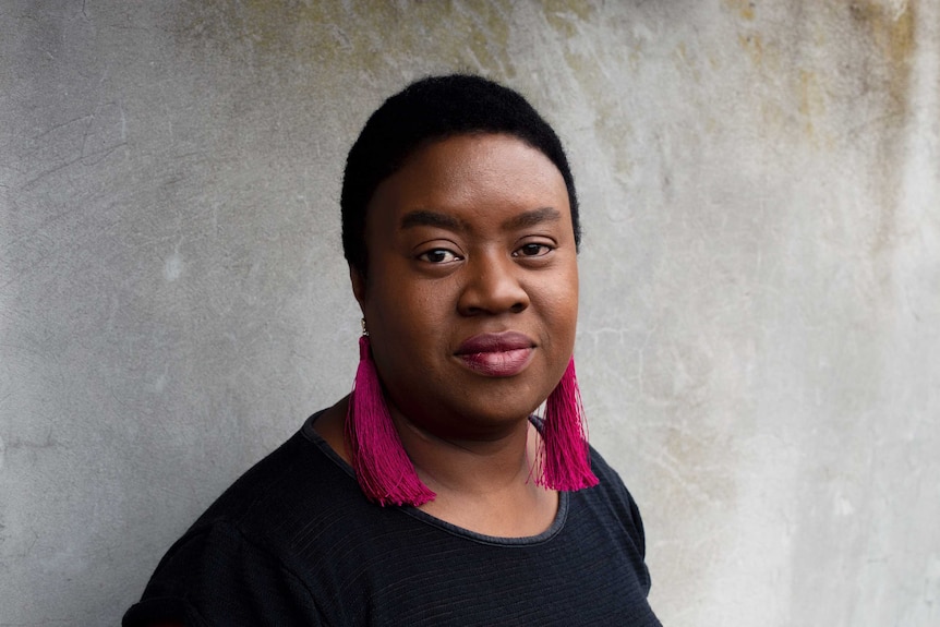 The writer Maxine Beneba Clarke standing in front of a cement wall, looking calmly determined