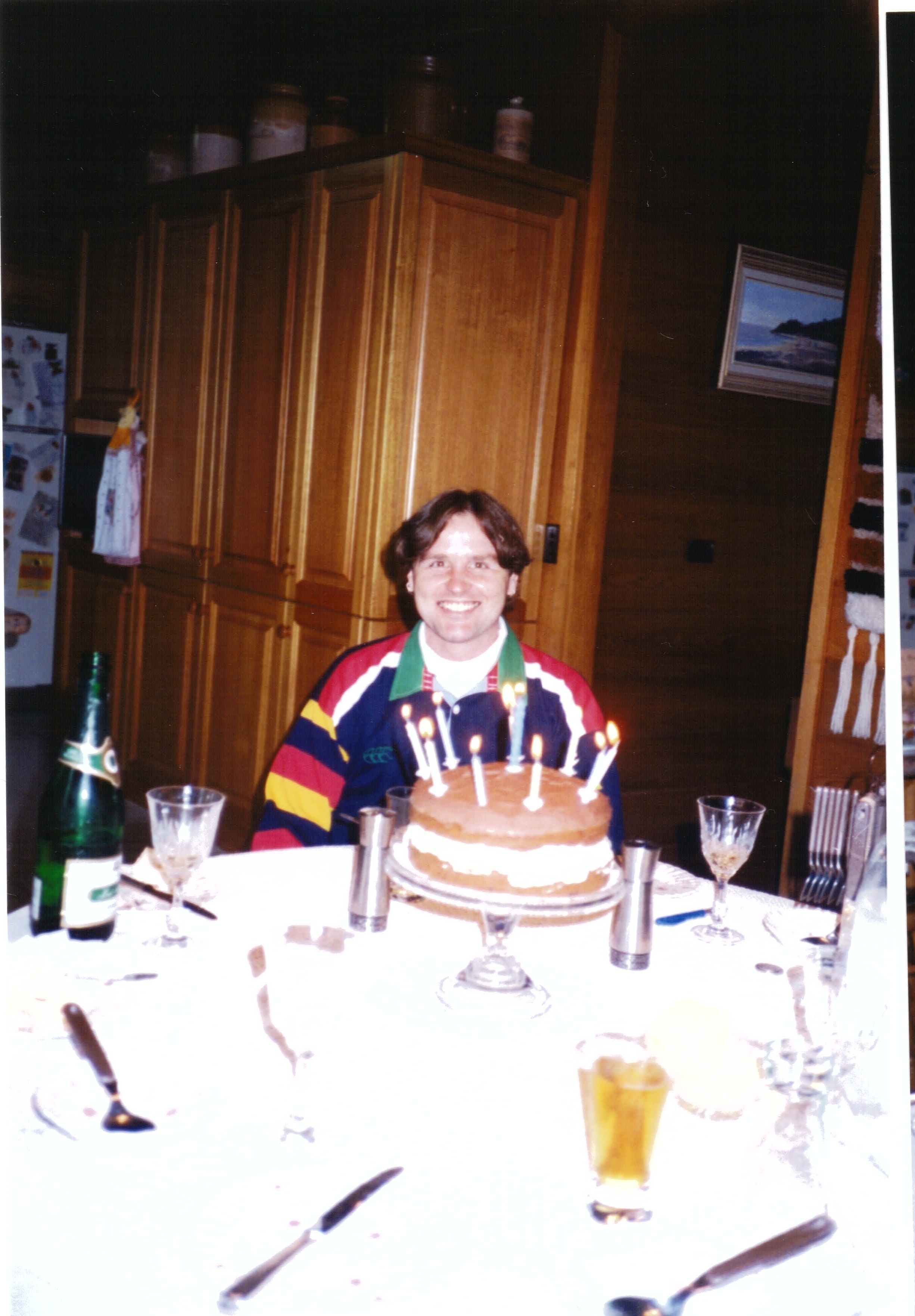 A man with brown hair sits at a table smiling. Before him is a birthday cake.