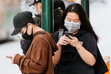 Two women in face masks at a Melbourne tram stop look at their phones.