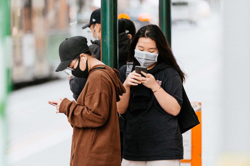 Two women in face masks at a Melbourne tram stop look at their phones.