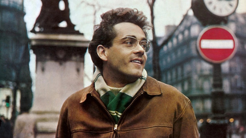 Michael Legrand in a brown coat with glasses walking through the streets of Paris
