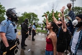 A police officer wearing a riot mask watches on as protesters raise their fists in the air.