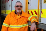 Volunteer firefighter Phil Penny stands in front of a Yarloop fire truck wearing an orange hi-vis jacket.