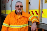 Volunteer firefighter Phil Penny stands in front of a Yarloop fire truck wearing an orange hi-vis jacket.