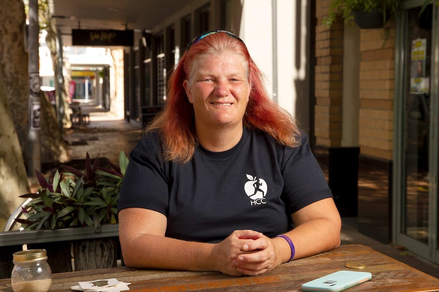 A woman with died red hair and a black T-shirt sits at a cafe bench in a street setting.