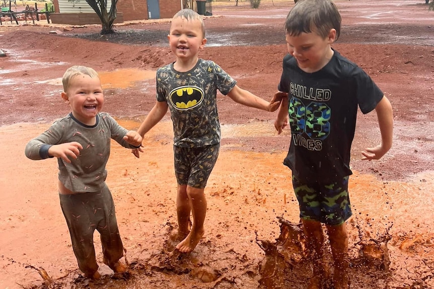Three kids jump in mud puddles at an outback station in rural SA,. They are smiling. 