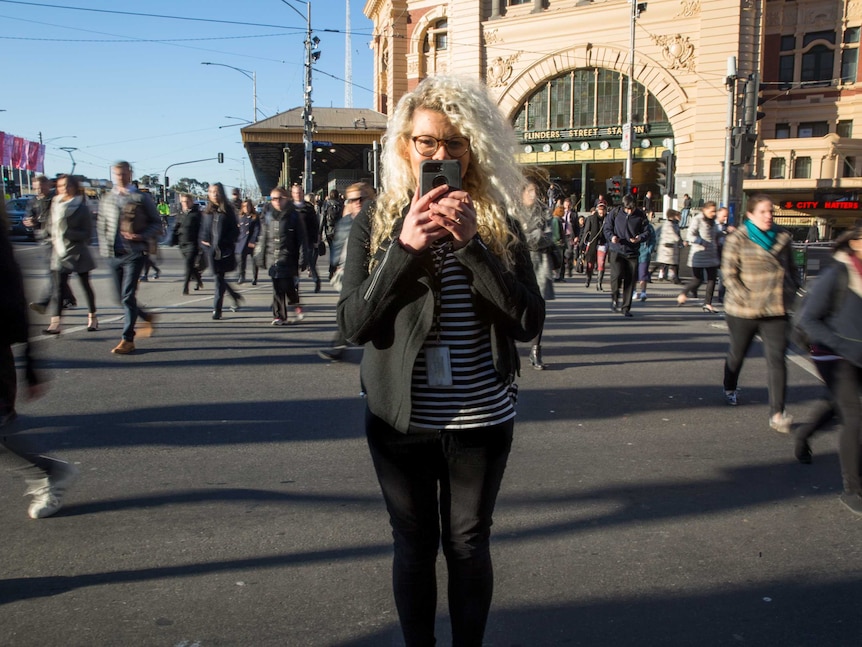 Margie Burin stands in the middle of Flinders Street crossing with a phone in front of her face as people walk by.