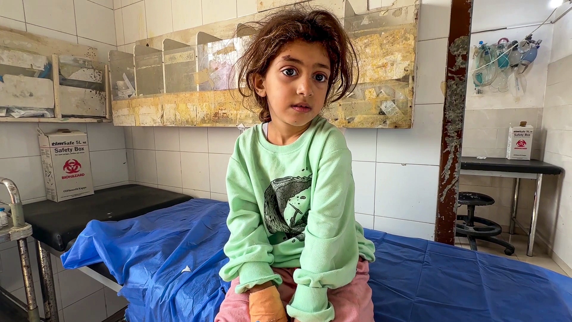 A small, injured girl sits on a hospital bed in a rundown room with a bandaged right hand.