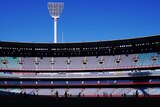 The afternoon sun hits the stands of an empty MCG stadium with players on the ground in silhouette.
