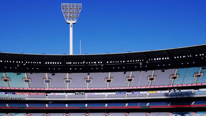 The afternoon sun hits the stands of an empty MCG stadium with players on the ground in silhouette.