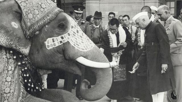 A black and white photo of the Dalai Lama and others with a decorated elephant in New Delhi.