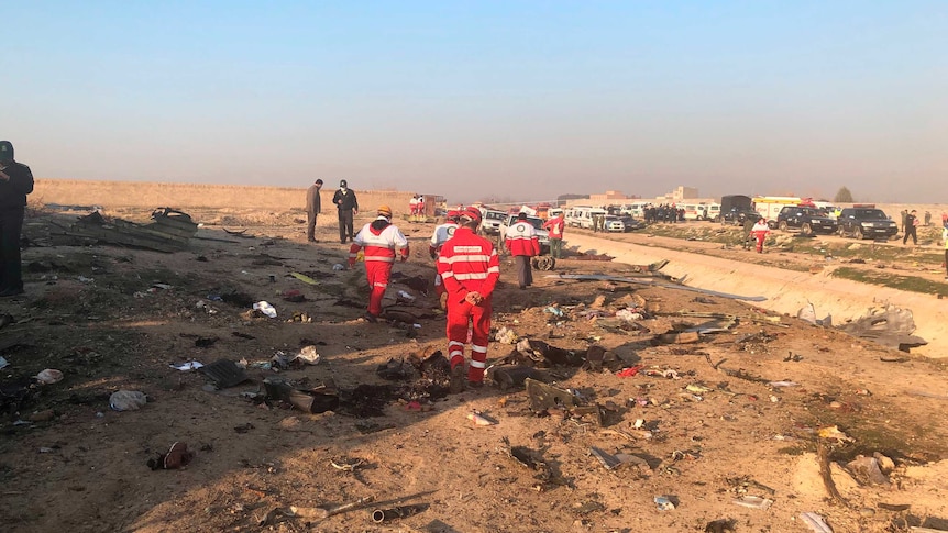 Rescue workers in red suits are seen looking over the wreckage of a plane. There are several emergency services vehicles.