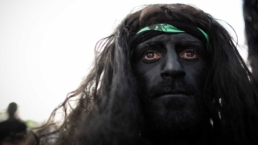 A man with long hair and a painted face peers out against a bright background.