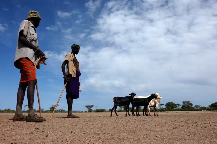 men stand on flat, dry dusty ground herding goats under a big blue sky with clouds