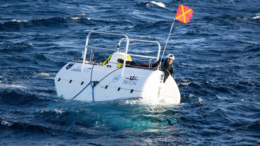 a man with a helmet aboard a submersible above the ocean with a red flag with a yellow cross on it