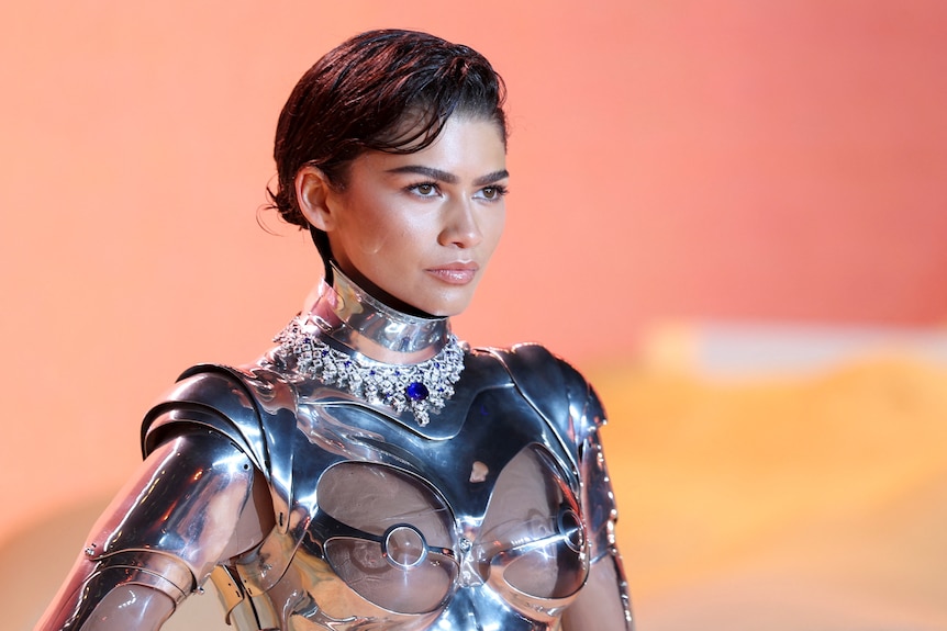 A close up of Zendaya against an orange backdrop wearing a metallic silver robot suit