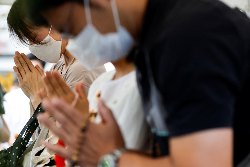 A line of people wearing masks with their head kneeling over their hands in prayer.