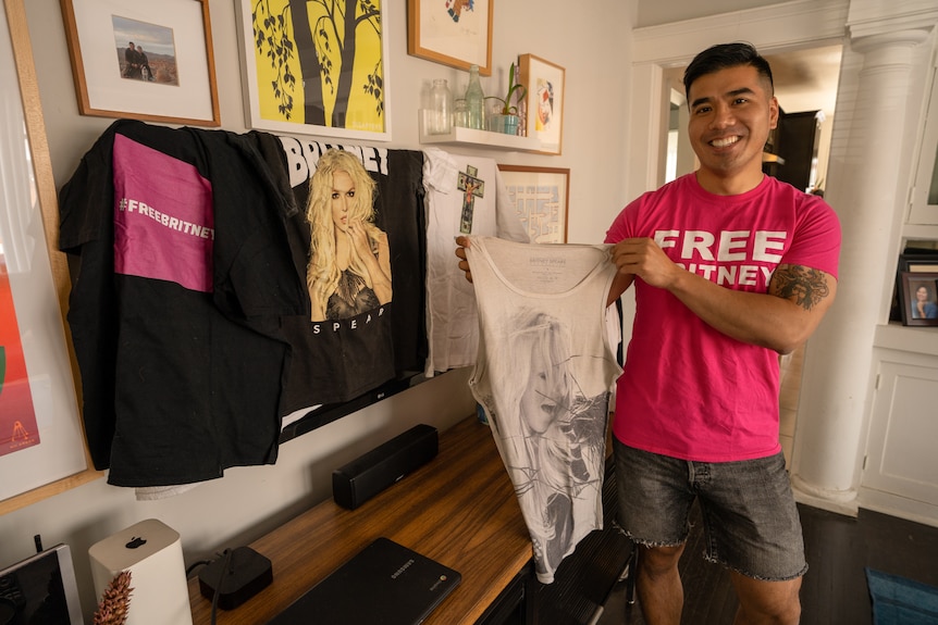 A man wears a pink shirt and holds up a tshirt of Britney Spears while standing with other shirts.