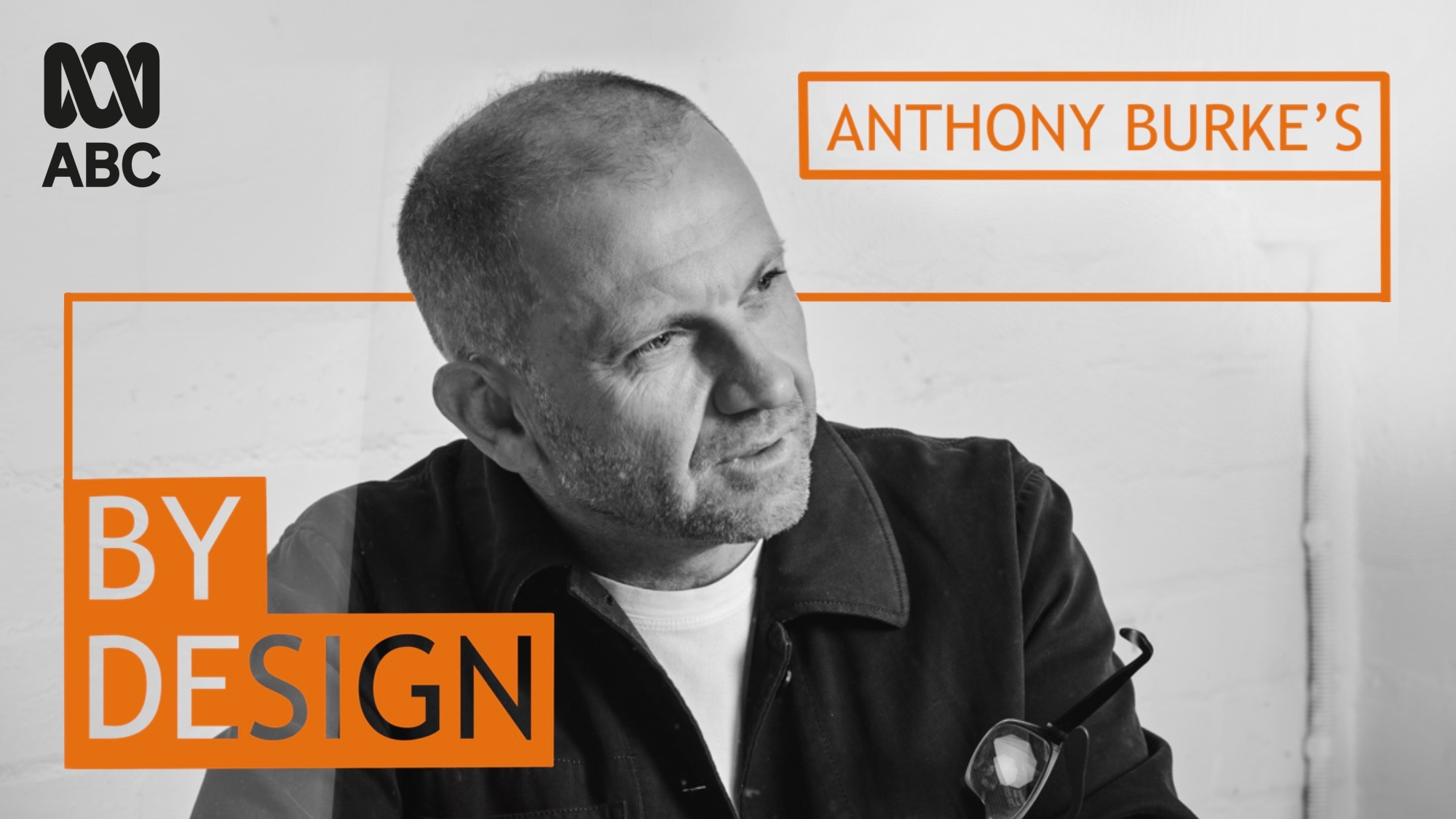 A man in black in white holding glasses looks to the side and the words Anthony Burke's By Design