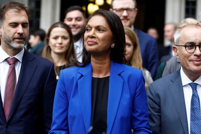 Gina Miller smiles with her mouth closed as she is flanked by numerous people. She wears a blue blazer and has long dark hair.