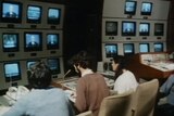 Grainy footage from 1979 of three people working in an old news studio with screens covering one wall.