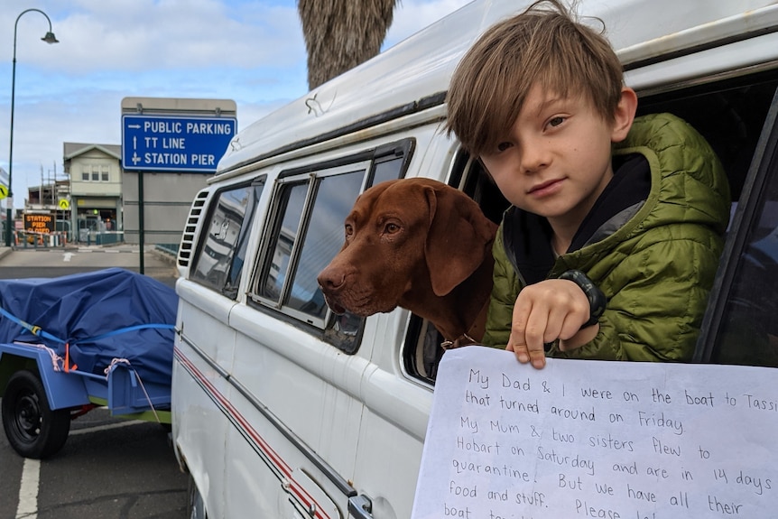 A little boy in green jacket holds white sign out of white van with brown dog in window behind him