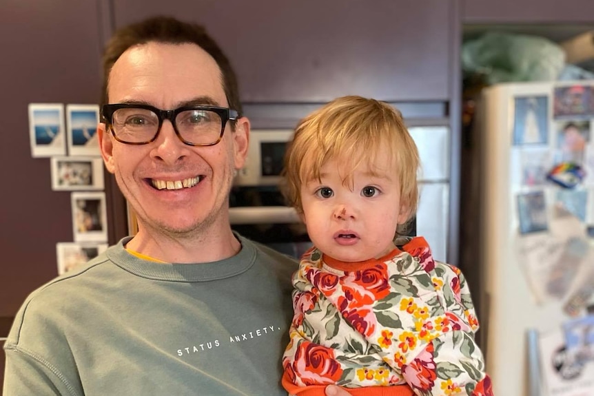 In a kitchen, a man with thick-rimmed glasses smiles widely as he holds a small boy in his arms.