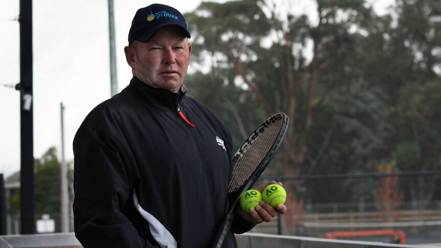 Graham Charlton holds a tennis racquet and balls overlooking tennis courts.