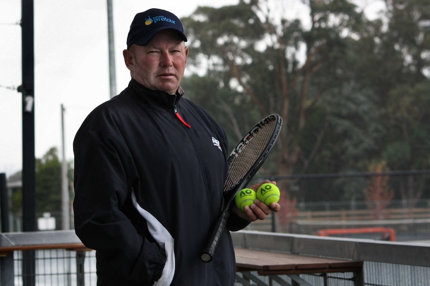 Graham Charlton holds a tennis racquet and balls overlooking tennis courts.