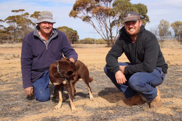 Max and Joel Lancaster kneel on the ground in a dry farm paddock with their dog Fred.