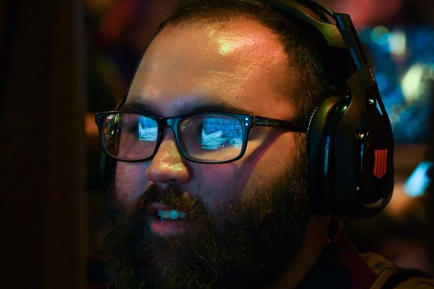 A computer screen can be seen in the reflection of a man's glasses. He is wearing headphones.