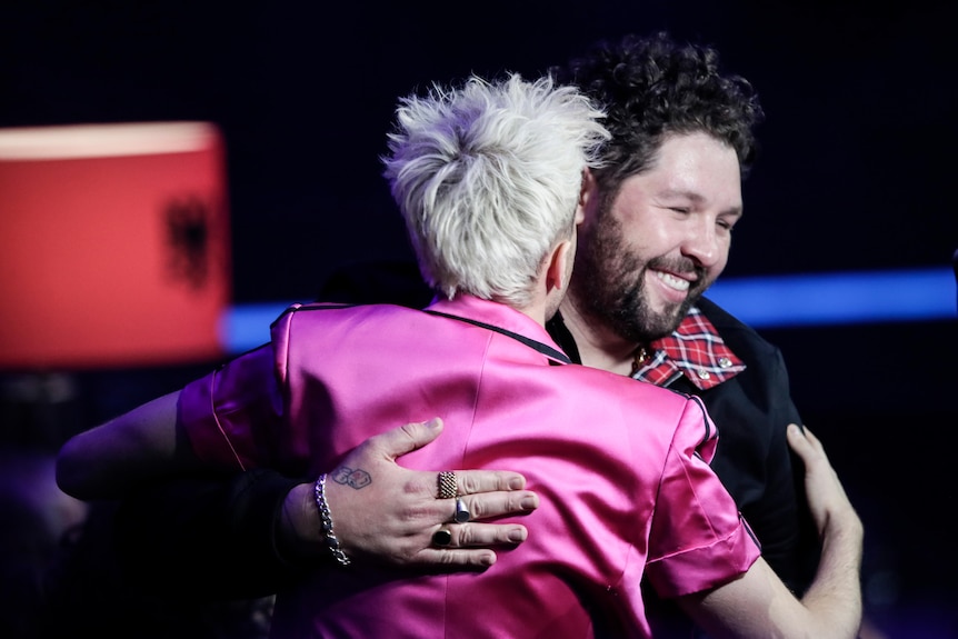 A bearded singer is hugged by a man in a pink jacket in the backstage area at the Eurovision Song Contest.