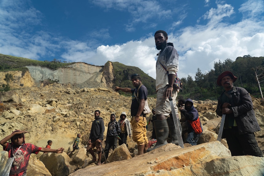 A group of mostly young men stand on top of a large pile of rubble by a sheer hillside, some of them holding machetes.